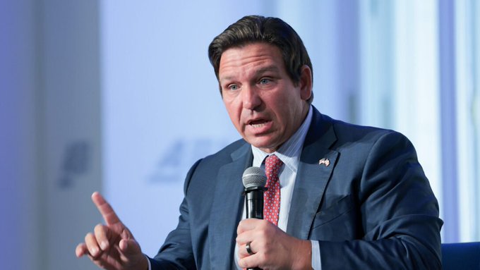 Florida Governor Ron DeSantis, wearing a dark suit and red tie, speaks into a microphone while gesturing with his hand during a public appearance or interview.