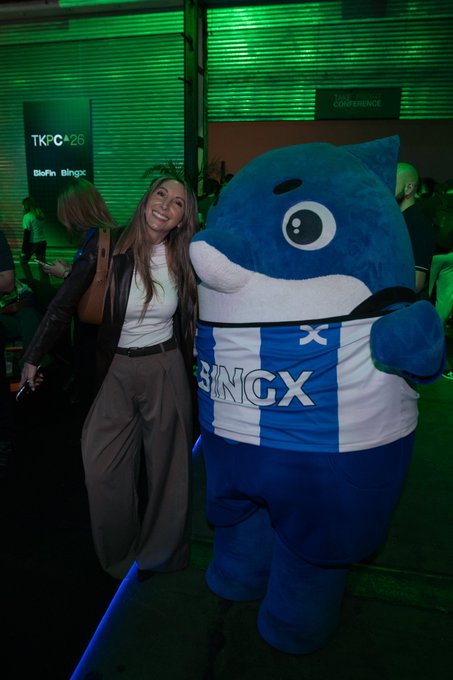 A woman posing with a large blue shark mascot wearing a BINGX jersey at the TKPC'26 conference.