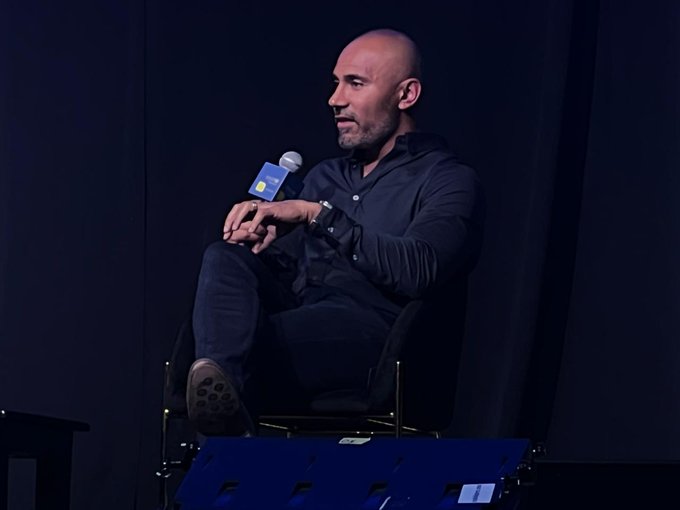 In this image, a bald man wearing a black shirt sits in a chair with his legs crossed while holding a microphone. The background shows a stage or podium set up for an event, possibly a conference or gathering related to stablecoins. The caption provided by the image suggests that Raja Chakravorti and Ximena Salgado are also present at this event.