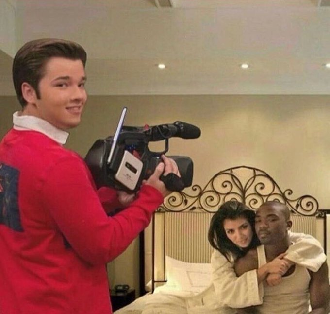 A vintage behind-the-scenes photo showing a cameraman in a red sweater filming a scene on a bed with an ornate metal headboard, where a man and woman in white robes are embracing.