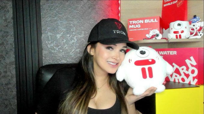 A smiling woman wearing a black "TRON" cap holds up a plush TRON mascot (BitBull) in front of a display featuring TRON-branded mugs and merchandise, visually reinforcing her bullish post about #TRX being added to the S&P Pantera Digital Asset Index.