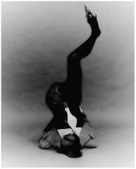A black and white photograph of a performer in a striking acrobatic pose, balancing on their head with legs extended vertically.