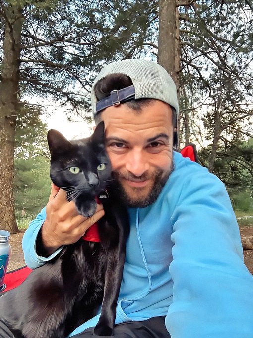 A selfie of a smiling bearded man in a blue hoodie and backwards cap, holding a black cat close to his face outdoors among trees.