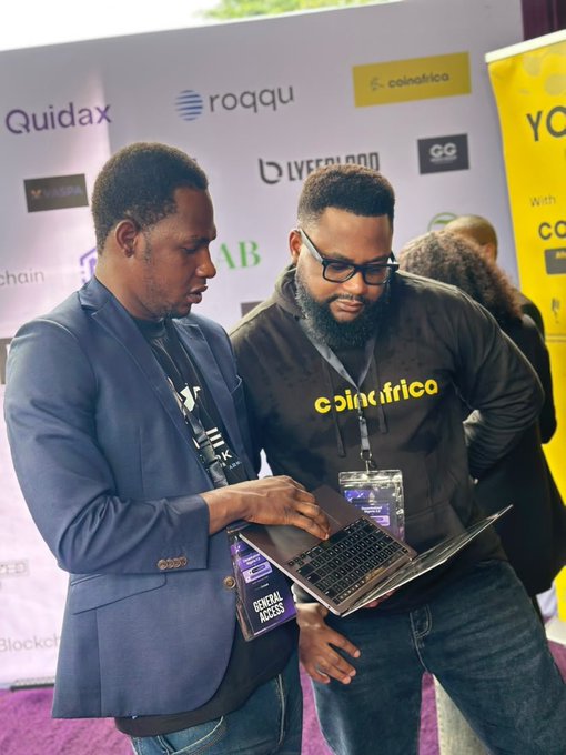 Two attendees at a crypto/blockchain conference—one in a "CoinAfrique" branded hoodie and the other in a blazer with a "General Access" badge—closely examine a laptop screen, likely introducing a newcomer to digital asset platforms like Roqqu, Lyfeblood, and Quidax displayed on the backdrop.