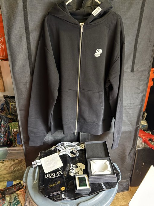 A black zip-up hoodie with a small white skull logo hangs on display above folded clothing items labeled 'LUCKY APE', with a price tag reading '12.8'.