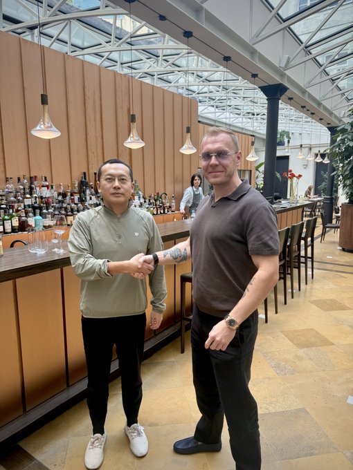 Two men shake hands in a modern, upscale bar or restaurant with a glass ceiling, as part of a post praising @star_okx for incorporating community user feedback into tangible perks.