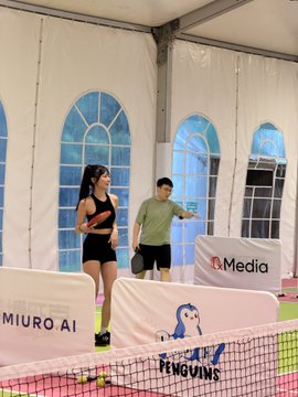 Two people stand on an indoor pickleball court under a white tent, with sponsor banners reading 'MIURO.AI', 'PENGUINS', and 'n.Media' displayed on the net.