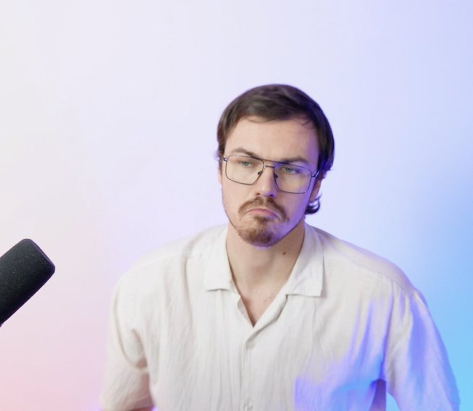 A man with glasses and a goatee makes a displeased expression while sitting in front of a microphone against a pink and blue lit background.