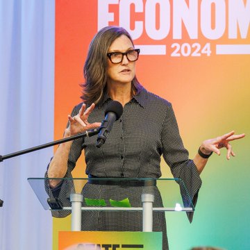 ARK Invest CEO Cathie Wood speaks at a podium during an "ECONOMY 2024" event, gesturing with her hands as she addresses the audience. The image accompanies her recent remarks praising Bitcoin as a revolutionary new monetary system and asset class with significant growth potential still ahead.