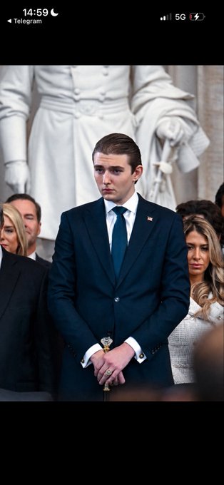 A screenshot from Telegram shows Barron Trump standing in a dark suit with an American flag pin, positioned in front of a white statue.