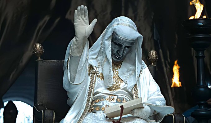 A pale, robed figure with a hood and a long beard sits on a throne, raising one white-gloved hand while holding a scroll in the other, with flames burning in the background.