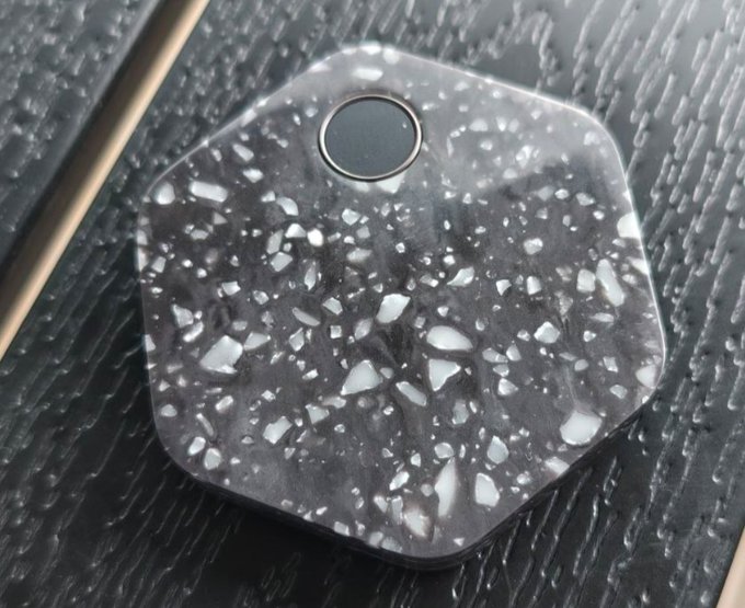 A hexagonal Bitkey hardware wallet with a distinctive terrazzo-style speckled black and white pattern rests on a textured dark surface, illustrating the post's point about each unit's unique hardware appearance. The single circular button and minimalist design emphasize the device's one-of-a-kind aesthetic, as no two Bitkeys share the exact same terrazzo pattern.