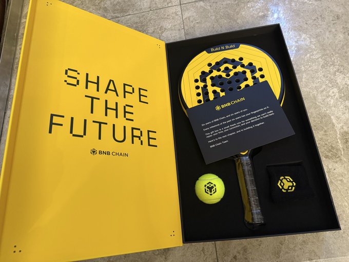 A BNB Chain branded padel set in a yellow box with the text 'SHAPE THE FUTURE', including a yellow padel racket, tennis ball, and sweatband with BNB Chain logos.