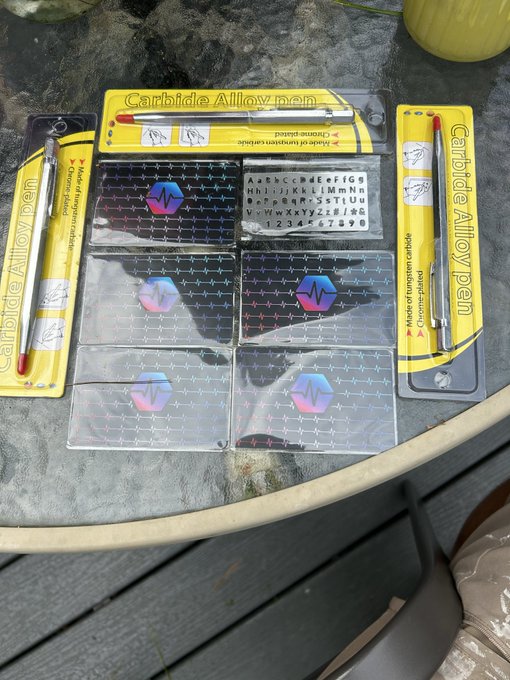 Several packaged 'Carbide Alloy pen' sets and clear plastic sheets containing metallic grid stencils with heart rate and waveform logos, laid out on a glass table.