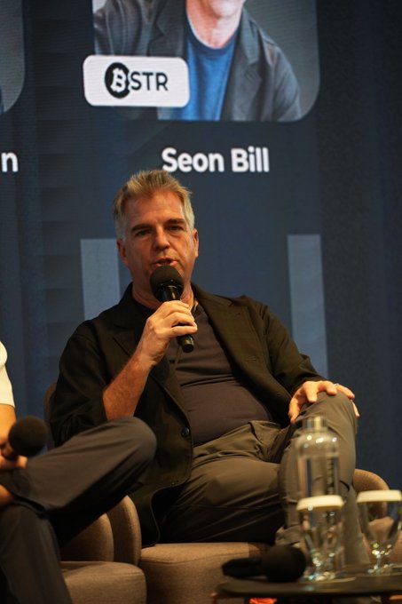Sean Bill, co-founder of Bitcoin Standard Treasury Company (BSTR), speaks into a microphone during a panel discussion, with a screen behind him displaying "BSTR" and his name "Seon Bill." The image accompanies commentary on his remarks about limited Bitcoin adoption among U.S. pension funds.