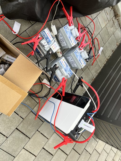 The image shows a collection of batteries, including four AAA batteries and two AAs, connected by red jumper cables. The batteries are placed on a brick sidewalk next to a cardboard box that has been opened. There is also a black bag nearby. The scene appears to be related to an off-grid project or experiment involving the use of these batteries for power supply.