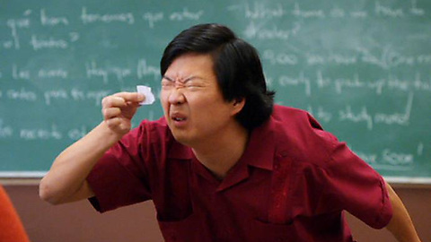 A man with shoulder-length black hair in a red shirt stands in front of a chalkboard covered in writing, dramatically squinting while holding up a small white object.