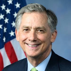 An official portrait of a smiling man with gray hair in a dark suit and green tie, standing in front of an American flag.
