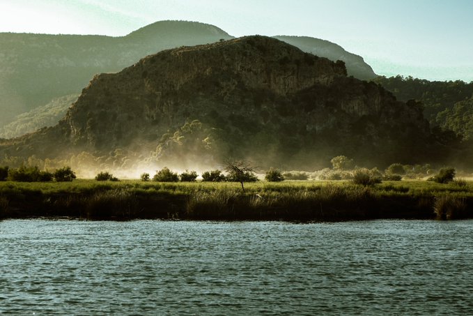 A scenic landscape featuring a large rocky hill or mountain rising behind misty grasslands and reeds, with calm water in the foreground under a hazy, atmospheric sky.