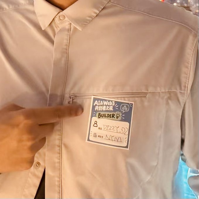 A close-up of a person pointing at a handwritten "AI & Web3" name tag on their shirt that reads "BUILDER" with alphanumeric codes.