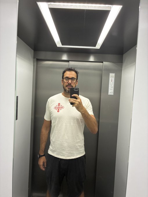 A man wearing glasses takes a mirror selfie inside an elevator, sporting a white t-shirt featuring a red diamond-shaped "Solana" logo and dark shorts. The image showcases the Solana Summit Serbia event merch the poster is proudly adding to their daily rotation.