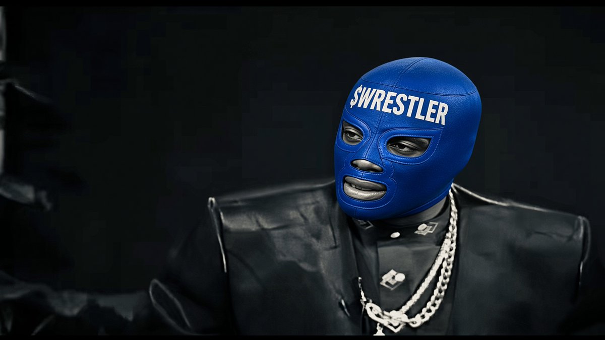 A person wearing a blue wrestling mask with '$WRESTLER' written in white text across the forehead, dressed in a black leather jacket with silver chains against a black background.