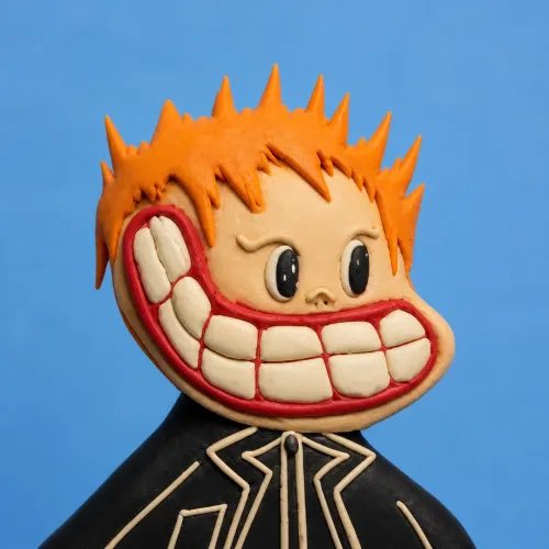 A claymation-style figure with spiky orange hair, an oversized grinning mouth with large white teeth, and a black collared jacket against a blue background.