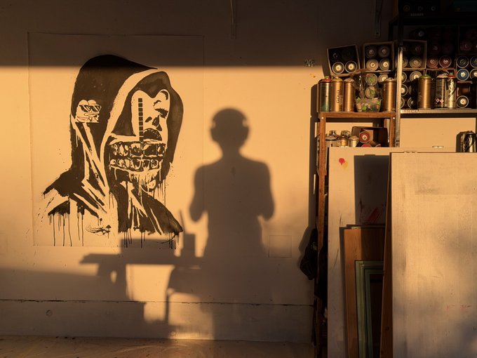 A large black-and-white graffiti mural of a hooded skeletal figure with prominent gold teeth is displayed on a wall in a sunlit studio, with the shadow of the photographer cast across the artwork next to shelves of spray paint cans.