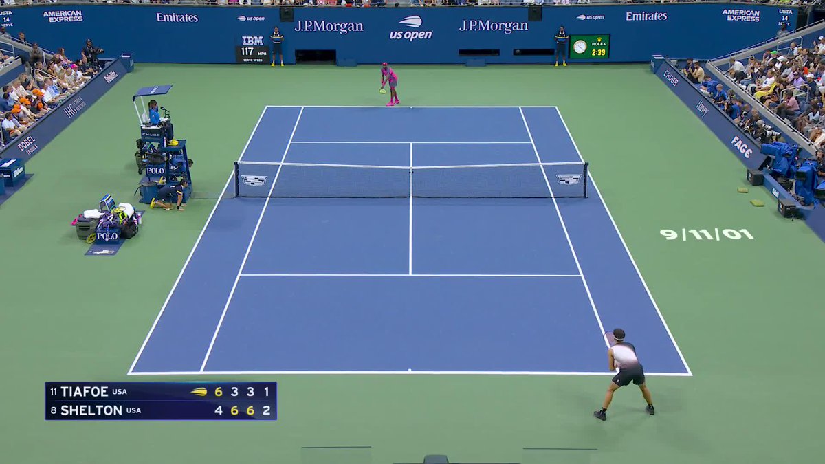 A tennis match between Frances Tiafoe (#11) and Ben Shelton (#8) at the US Open, with the scoreboard showing Tiafoe leading 6-4, 3-6, 3-6, 1-2. Shelton, positioned at the baseline on the right side of the court, appears to be preparing to execute the cross-court shot mentioned in the post, while Tiafoe stands ready at the opposite baseline in his pink outfit.