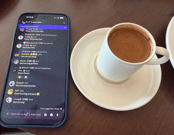 A smartphone displaying a Telegram 'main-chat' group with messages like 'Happy Wednesday guys!' and 'Gemmorning everyone' sits on a wooden table next to a white cup of espresso on a saucer.