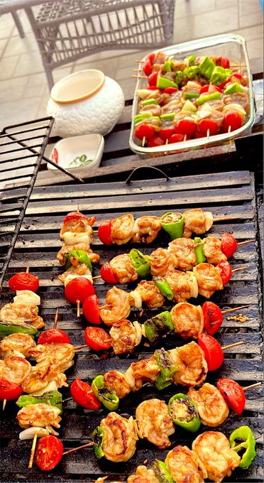 Shrimp and vegetable skewers sizzle on a charcoal grill while a tray of raw, marinated shrimp-and-pepper kebabs awaits its turn to be cooked, complementing the post's carefree "let's eat shrimp" attitude amid market turbulence.