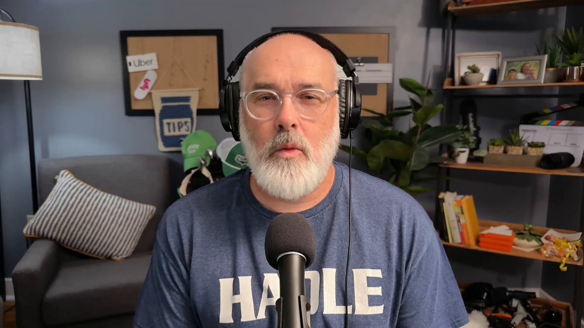 urns, plants, and a lamp are arranged on a shelf against a blue wall, with a man wearing glasses and a microphone headset in the foreground.
