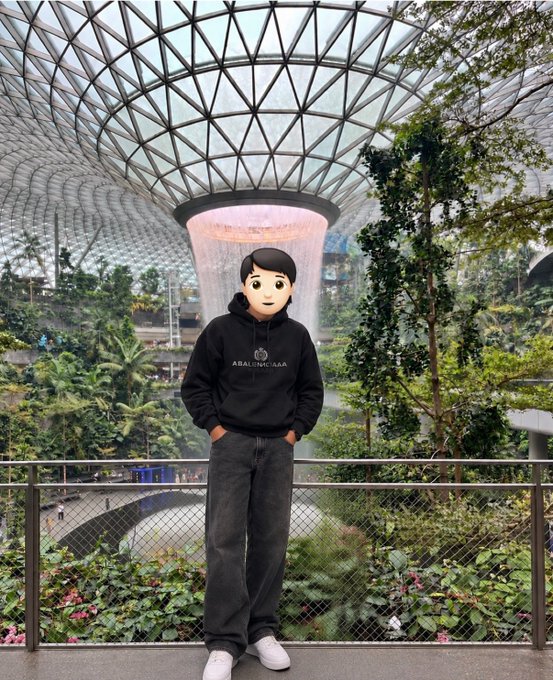 A person with an emoji face stands on a balcony at Jewel Changi Airport's indoor waterfall, wearing a black hoodie with "ARALUNA" branding. The location serves as a fitting farewell spot for Token 2049 attendees, signaling a wrap-up to the conference with an invitation to reconnect.