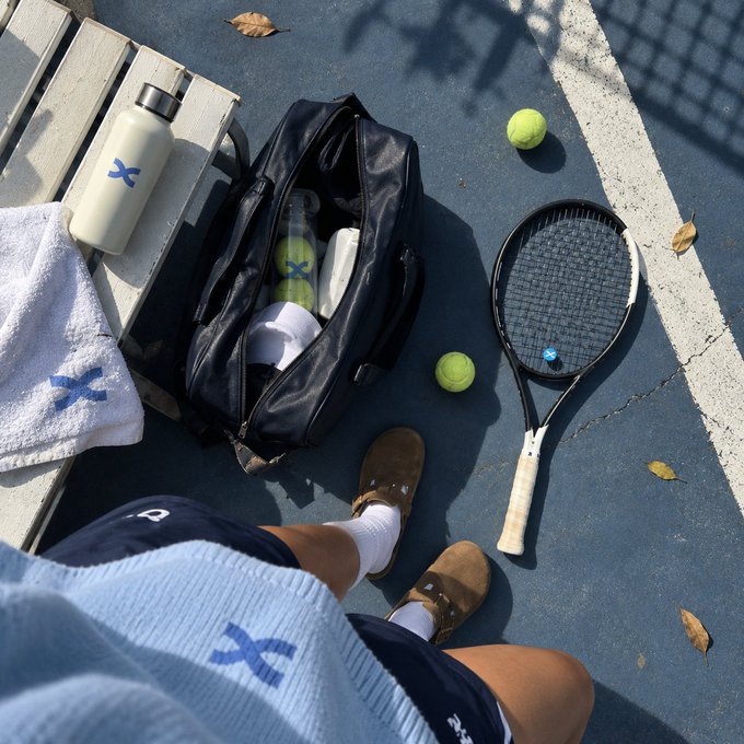 A first-person view of a tennis player on a blue court, with their bag, racket, balls, and a white water bottle marked with a blue "X" at their feet, complementing the post's note that the court itself is blue.