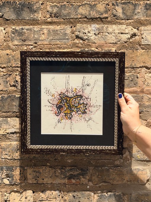A hand with dark blue nail polish holds up a framed pixel art piece resembling a Magikarp Pokémon against a rustic exposed brick wall, with the ornate distressed frame adding a striking contrast to the retro, 8-bit style artwork inside.