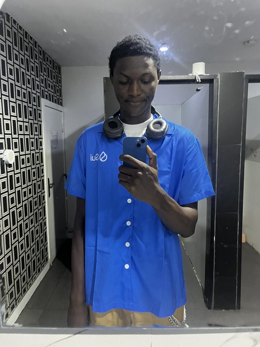 A young man taking a mirror selfie while wearing a blue IU (Indiana University) scrub top and headphones around his neck, standing in what appears to be a bathroom.