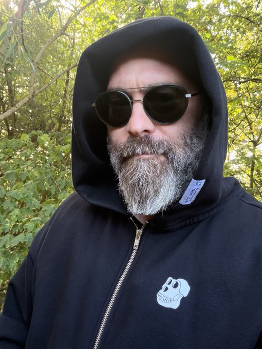 A close-up selfie of a bearded man wearing round sunglasses and a black zip-up hoodie with a skull design, standing outdoors near green foliage.
