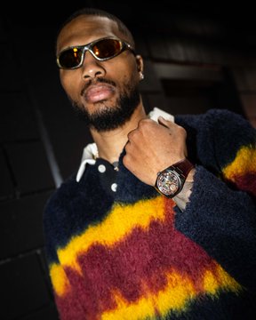 A man poses in sunglasses and a fuzzy multicolored sweater while displaying a large wristwatch, matching the post’s focus on the Satoshi x Oakley shades and Dame x TISSOT watch. No readable text is visible in the image.