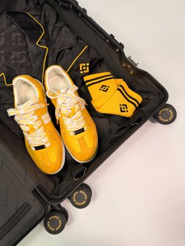 A pair of yellow sneakers sits inside an open black suitcase alongside a folded yellow shirt, all branded with a distinctive logo.