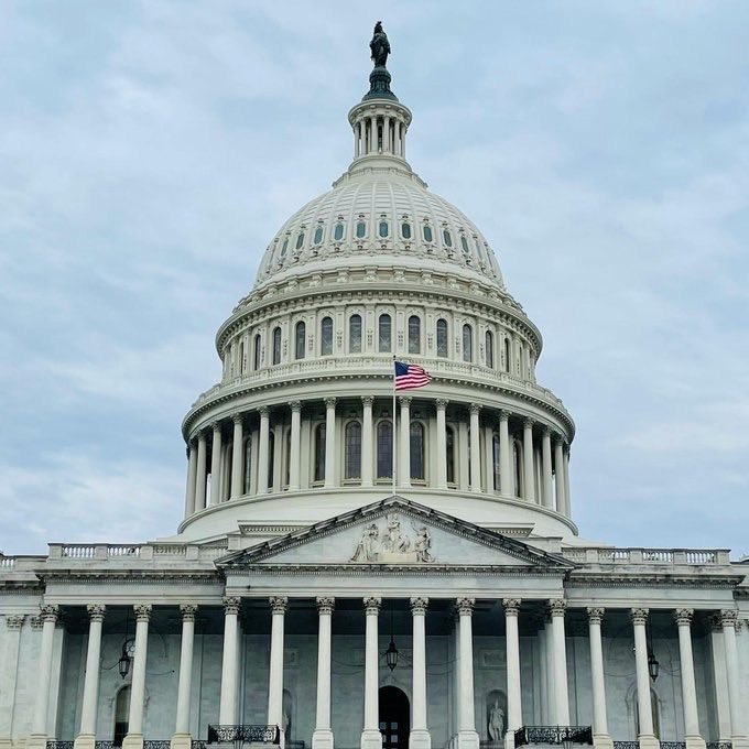 A photograph of the United States Capitol Building dome with an American flag flying in front of it against a cloudy sky.