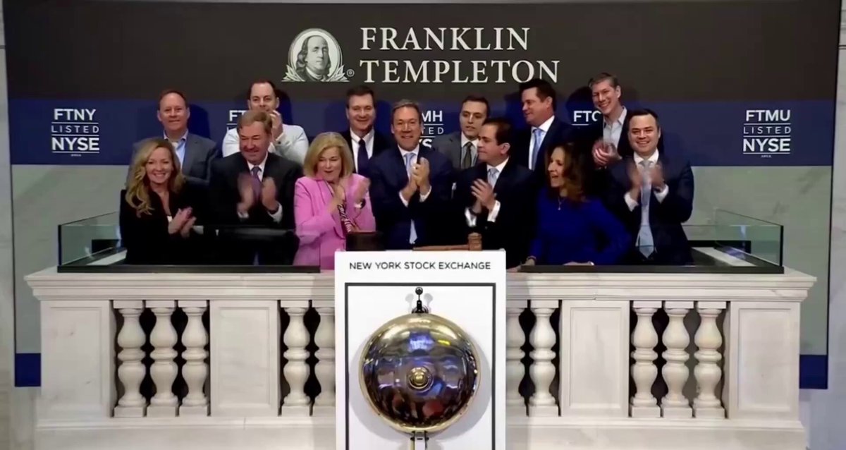 The image features a group of people standing together, with some of them clapping their hands. They are gathered around a large clock that has a gold face and white numbers. The clock is mounted on a wall next to a podium, which also has a gold face and white numbers. In the background, there is a sign for Franklin Templeton, indicating that this gathering might be related to an event or celebration involving Franklin Templeton ETFs.
