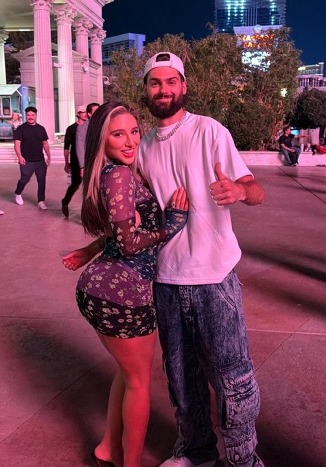A couple poses together at night outside what appears to be a venue with classical columns, the man giving a thumbs-up while wearing a white cap and tee with baggy jeans, and the woman in a floral dress smiling beside him—capturing the social, nightlife energy that fits the "SOLANA SUMMER" vibe of crypto's vibrant party scene.