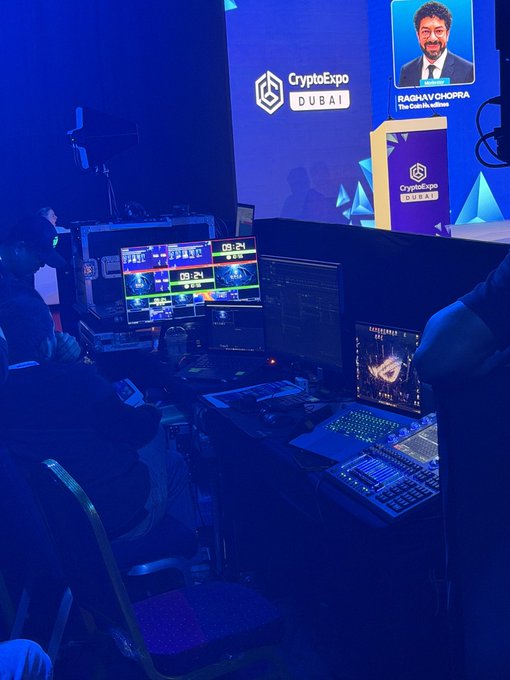 Behind-the-scenes view of a production control room at Crypto Expo Dubai, where a crew operates multiple monitors, a video switcher, and laptops under blue lighting while the event displays "CryptoExpo Dubai" branding and a portrait of host Raghav Chopra on the main screen.