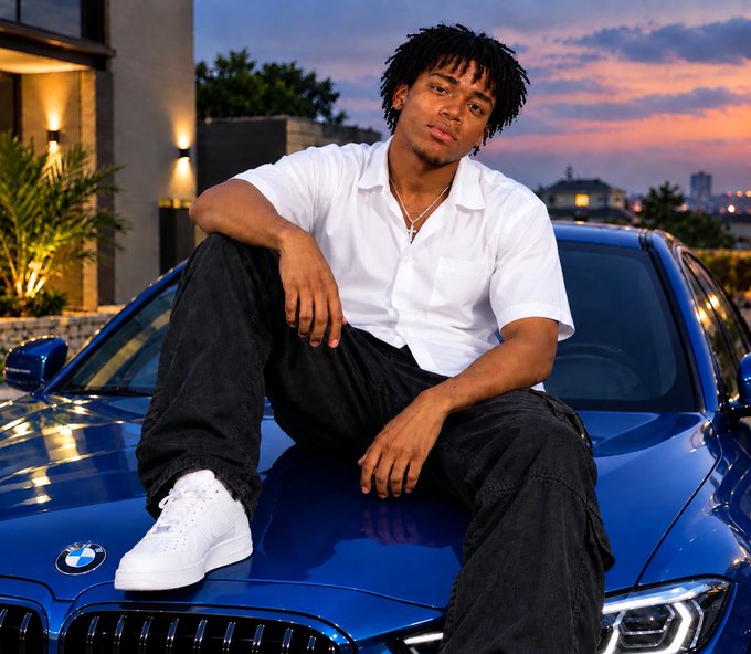 A young man in a white shirt and black pants sits on the hood of a blue BMW at sunset, posing for a stylish portrait photo.