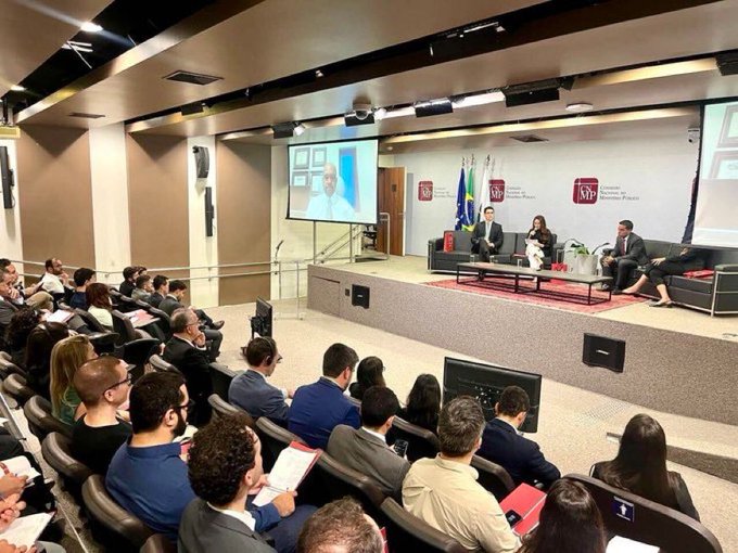 An audience seated in an auditorium watching a panel discussion on a stage with an 'MPF' (Ministério Público Federal) backdrop, featuring speakers and a video call displayed on a projection screen.