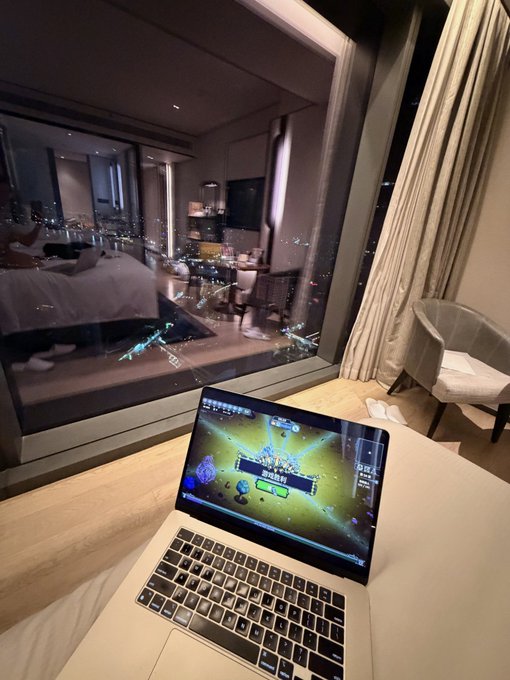 An open laptop displaying a fantasy MMORPG game rests on a bed in a high-rise hotel room, with a nighttime city skyline visible through the large window in the background.