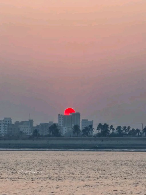 A sunset over a coastal city skyline with a red sun partially hidden behind buildings and palm trees, with the text "BeautyofBangladesh" visible in the lower left corner.