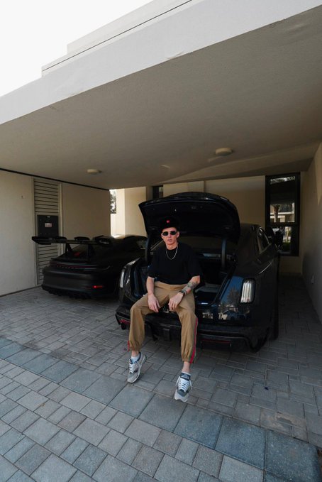 A person wearing sunglasses and a black cap sits in the open trunk of a black Porsche, with another black Porsche parked in the garage behind them.