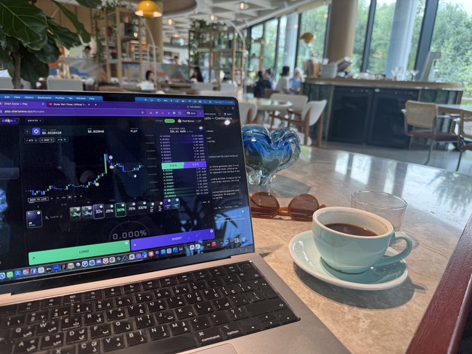 A MacBook laptop on a table at a café displaying a trading/charting application (appears to be a crypto or stock trading platform), with a cup of espresso, sunglasses, and a heart-shaped glass decoration beside it.