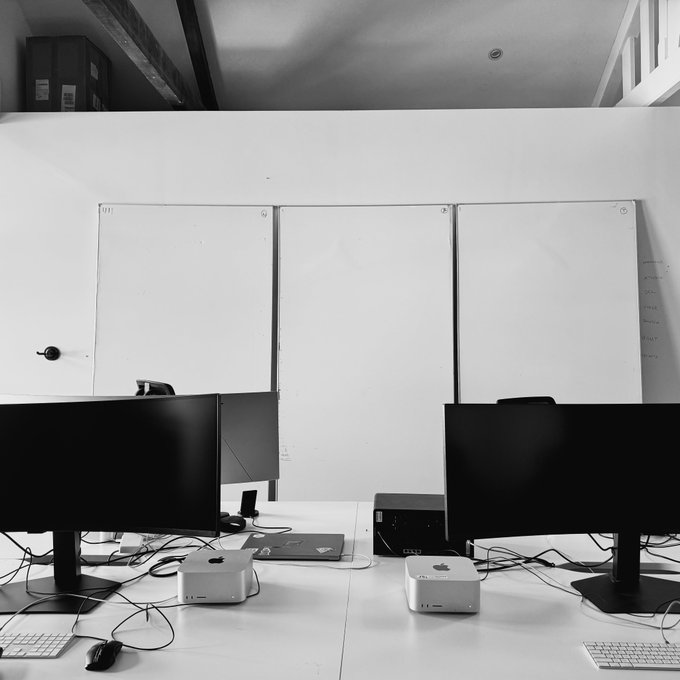 A black and white photo of an office or classroom with two computer setups featuring Apple monitors and Mac Mini computers in front of whiteboards.